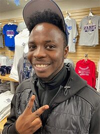 Image of a male wearing a hat, making a hand gesture while standing in front of a retail clothing display. This image would not be accepted for submission.