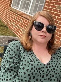 Image of a female wearing sunglasses while sitting outside with a brick wall background. This image would not be accepted for submission.